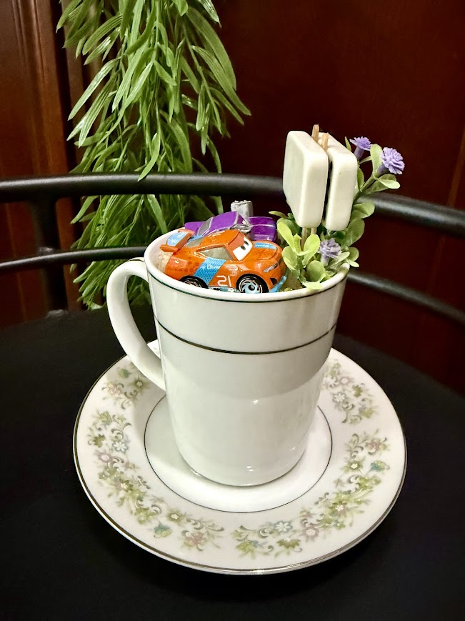 Toy racecar in a mug garden filled with sand, florals, and faux greenery.