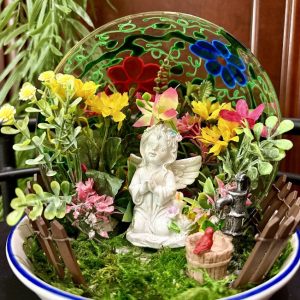 Heavenly-Angel-Dish-Garden Porcelain angel surrounded by colorful flowers and greenery in a shallow dish with a stained-glass backdrop.