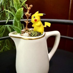 Pikachu-Forest-Mug-Garden-Craft Pikachu figurine standing next to faux plants inside a white pitcher, creating a playful garden.
