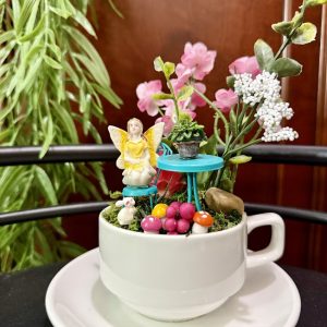 Sunshine-Fairy-Cafe-Cup Yellow fairy and miniature cafe table with flowers in a white teacup garden setting.
