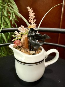 Ronin figurine in a white pitcher with flowing cape and decorative plants and stones.