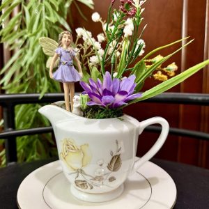 Purple fairy figurine in a floral creamer surrounded by faux flowers and greenery.