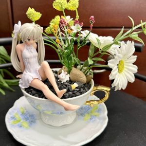 A whimsical anime girl figurine in a white dress sits in a floral teacup filled with black gravel, faux daisies, and colorful garden decor, including a bunny and pebbles.