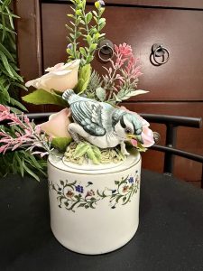 Bluebird figurine sitting among faux blush roses and greenery atop a vintage floral ceramic jar.
