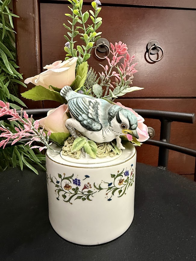 Bluebird figurine sitting among faux blush roses and greenery atop a vintage floral ceramic jar.