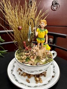 A boy figurine with a yellow-green outfit stands beside a dog, holding a fishing pole with a fish, surrounded by natural moss and earthy decor in a teacup.