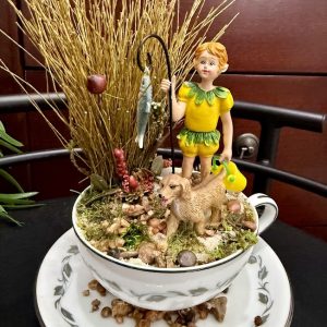 boy-and-dog-teacup-fishing-gnome-garden-decor A boy figurine with a yellow-green outfit stands beside a dog, holding a fishing pole with a fish, surrounded by natural moss and earthy decor in a teacup.