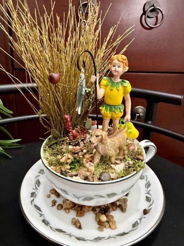 A boy figurine with a yellow-green outfit stands beside a dog, holding a fishing pole with a fish, surrounded by natural moss and earthy decor in a teacup.
