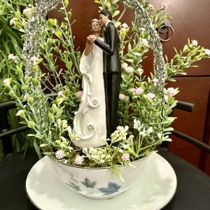 bride-and-groom-floral-wedding-keepsake-cup Figurine of a bride and groom dancing under a glittery arch, nestled in faux greenery and pink flowers, inside a floral teacup and saucer