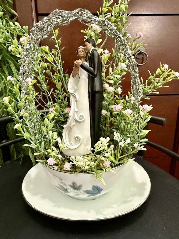 Figurine of a bride and groom dancing under a glittery arch, nestled in faux greenery and pink flowers, inside a floral teacup and saucer
