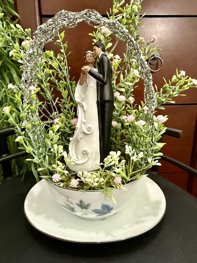 Figurine of a bride and groom dancing under a glittery arch, nestled in faux greenery and pink flowers, inside a floral teacup and saucer
