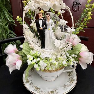 bride-and-groom-teacup-wedding-keepsake-floral-decor A bride and groom cake topper figurine stands inside a floral teacup decorated with white lace, bows, and pastel pink and green flowers.