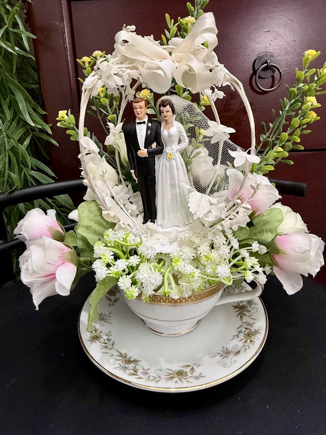 A bride and groom cake topper figurine stands inside a floral teacup decorated with white lace, bows, and pastel pink and green flowers.