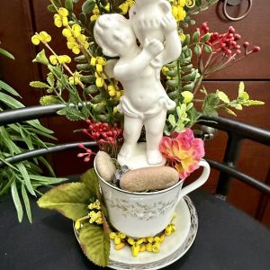 cherub-statue-floral-teacup-garden-romantic-decor A vintage teacup holds a white cherub statue surrounded by red and yellow faux flowers, greenery, and small stones, creating a romantic display.