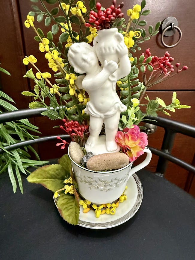 A vintage teacup holds a white cherub statue surrounded by red and yellow faux flowers, greenery, and small stones, creating a romantic display.