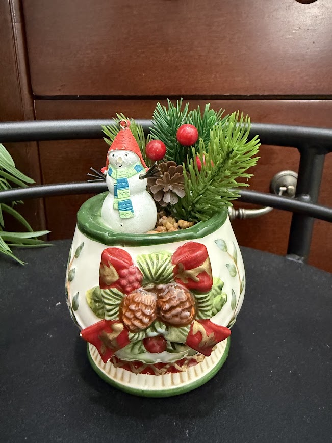 A holiday-themed mug with pinecone and ribbon decorations holds a joyful snowman figurine, pine sprigs, berries, and mini pinecones.