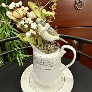 cream-bird-floral-pitcher-centerpiece-cottagecore-decor Cream-colored bird figurine with a floral necklace perched in a vintage floral pitcher with beige and white faux flowers.