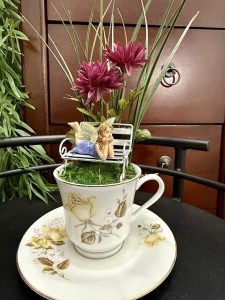 Whimsical fairy figurine lounging on a bench above moss, with purple flowers and greenery in a floral teacup and saucer.