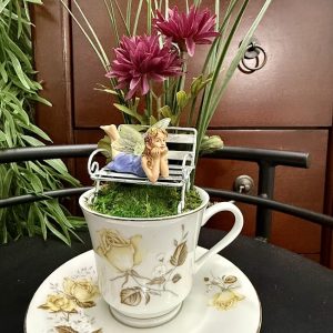 fairy-garden-bench-purple-flowers-teacup Whimsical fairy figurine lounging on a bench above moss, with purple flowers and greenery in a floral teacup and saucer.