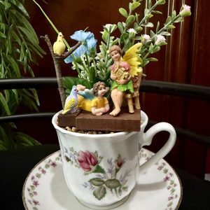 A floral teacup filled with a miniature fairy garden scene featuring two colorful fairies, a yellow bird on a twig perch, tiny flowers, and lush greenery. The cup sits on a matching saucer with pink and green floral detailing.