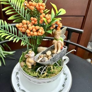 A fairy sits in a rustic wheelbarrow inside a teacup garden, accompanied by a bunny and orange berries with lush greenery.