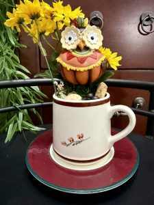 A fall-themed mug featuring a large pumpkin-colored owl figurine, surrounded by yellow flowers, berries, and rustic accents.