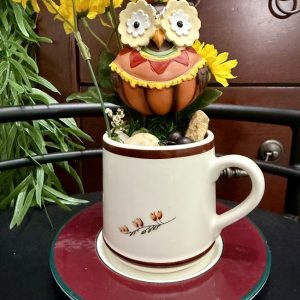 fall-owl-pumpkin-teacup-decor-autumn-mug-garden A fall-themed mug featuring a large pumpkin-colored owl figurine, surrounded by yellow flowers, berries, and rustic accents.