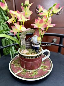 A teacup scene with a gnome figurine standing in front of a stone gate, surrounded by pink and yellow flowers, moss, and whimsical charm.