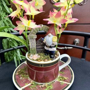 gnome-fairy-garden-teacup-with-gate-and-flowers A teacup scene with a gnome figurine standing in front of a stone gate, surrounded by pink and yellow flowers, moss, and whimsical charm.