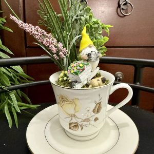 gnome-flower-cart-teacup-miniature-garden-decor A yellow-hatted gnome stands beside a white flower cart in a teacup garden, surrounded by greenery and delicate faux flowers.
