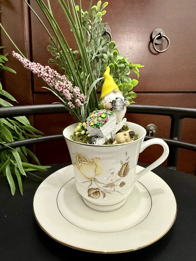A yellow-hatted gnome stands beside a white flower cart in a teacup garden, surrounded by greenery and delicate faux flowers.