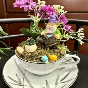 gnome-garden-teacup-purple-flowers-succulent-decor A cheerful garden gnome in a purple hat holds a watering can among bright pink flowers, moss, and succulents in a black and white teacup.