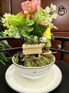 A gnome with a yellow hat tends to a wooden flower cart in a mossy teacup garden, watched by a small black dog