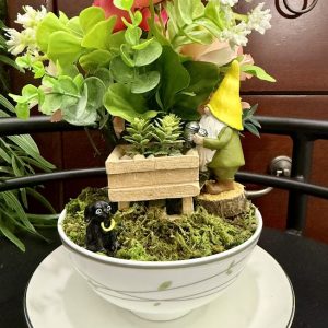 gnome-with-flower-cart-and-dog-teacup-garden-decor A gnome with a yellow hat tends to a wooden flower cart in a mossy teacup garden, watched by a small black dog