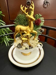 Golden reindeer figurines, including a stag and fawn, arranged on moss in a teacup with evergreen and pinecone accents.