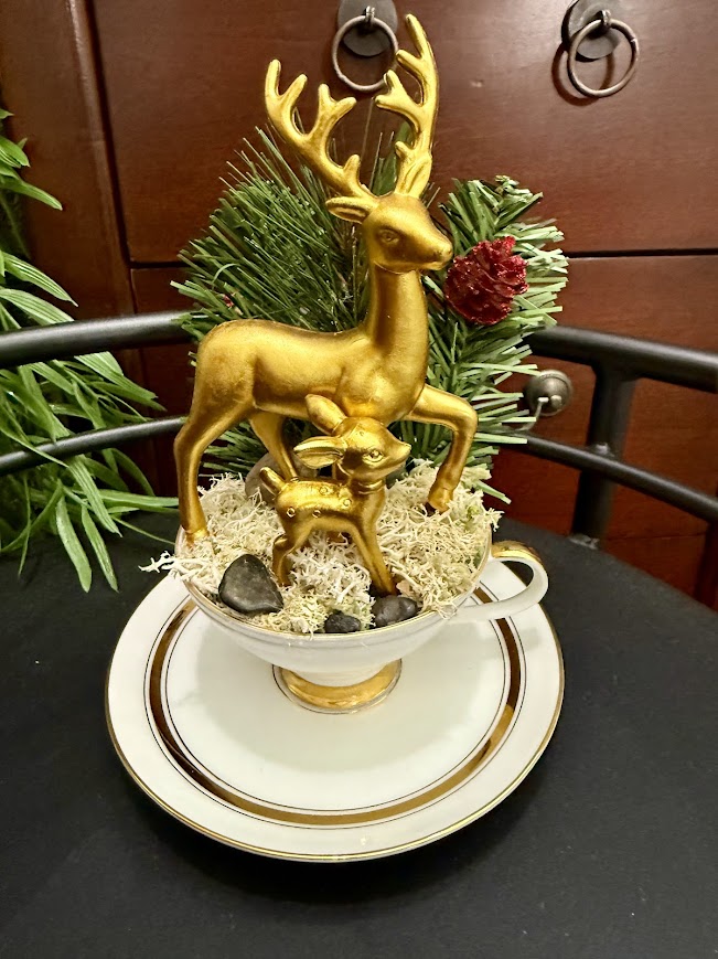 Golden reindeer figurines, including a stag and fawn, arranged on moss in a teacup with evergreen and pinecone accents.