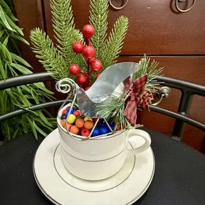 holiday-sleigh-candy-decor-white-teacup Miniature metal sleigh filled with colorful candies, pine sprigs, berries, and plaid ribbon, set inside a white teacup with silver rim.