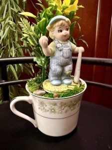 A whimsical boy figurine in blue overalls and a cap holding a baseball bat stands atop a floral teacup filled with faux moss, small stones, and bright yellow flowers. The teacup has a soft green and cream pattern and is placed on a dark surface.