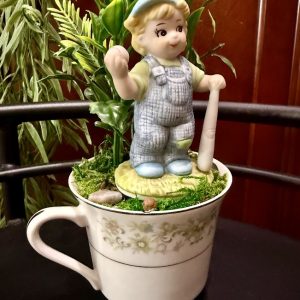 A whimsical boy figurine in blue overalls and a cap holding a baseball bat stands atop a floral teacup filled with faux moss, small stones, and bright yellow flowers. The teacup has a soft green and cream pattern and is placed on a dark surface.