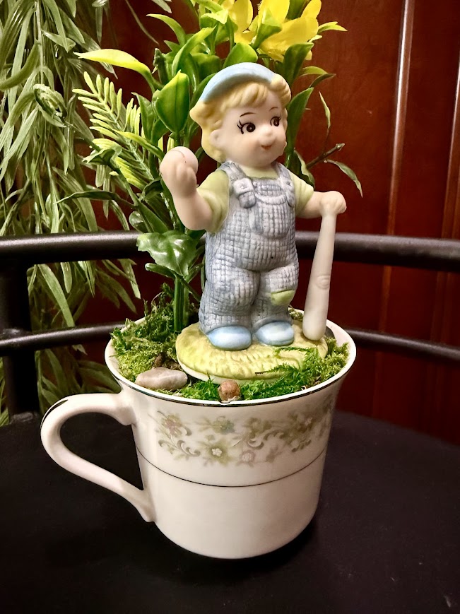A whimsical boy figurine in blue overalls and a cap holding a baseball bat stands atop a floral teacup filled with faux moss, small stones, and bright yellow flowers. The teacup has a soft green and cream pattern and is placed on a dark surface.