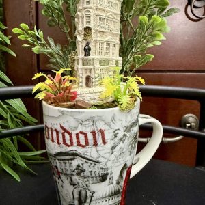 london-themed-mug-miniature-building-garden-decor A tall, historic-style miniature building rises from a โLondonโ themed mug decorated with city landmarks and surrounded by faux plants and flowers.