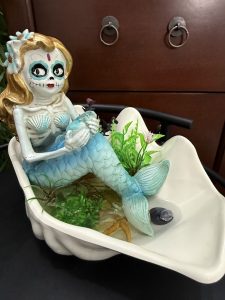 A blue-tailed mermaid with a Day of the Dead face rests in a white gravy boat filled with faux seaweed, a starfish, and decorative water elements.