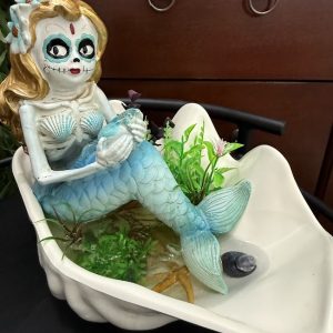 mermaid-day-of-the-dead-gravy-boat-ocean-fairy-decor A blue-tailed mermaid with a Day of the Dead face rests in a white gravy boat filled with faux seaweed, a starfish, and decorative water elements.