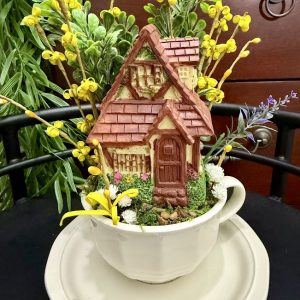 miniature-cottage-teacup-garden-house-fairy-decor A miniature cottage with a red roof nestled inside a teacup, surrounded by yellow flowers and greenery, resembling a storybook home.
