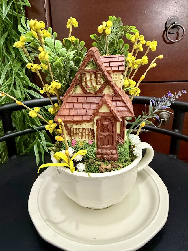 A miniature cottage with a red roof nestled inside a teacup, surrounded by yellow flowers and greenery, resembling a storybook home.