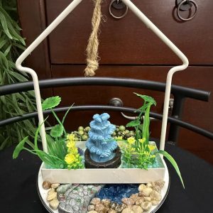 miniature-zen-garden-hanging-decor-water-feature A hanging ceramic tray garden features a miniature blue fountain surrounded by faux plants, pebbles, and decorative stones, evoking a peaceful zen retreat.