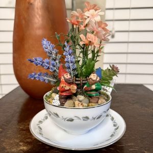 musical-gnomes-teacup-garden-lavender-decor Two colorful gnomes play instruments in a floral teacup filled with lavender, greenery, and river stones, creating a joyful garden scene.