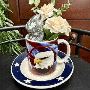 patriotic-military-mug-eagle-flower-decor American flag-themed mug featuring a bald eagle, filled with artificial roses, greenery, and a miniature digital camouflage military cap.