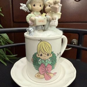 precious-moments-christmas-mug-miniature-noel-decor A Christmas-themed Precious Moments mug features two festive figurines, tiny gifts, and soft snowy fluff, creating a heartwarming holiday display.