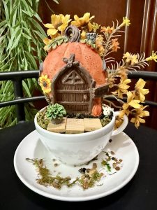 A pumpkin-shaped fairy house with a wooden door, sunflowers, and succulents sits atop a white teacup. Yellow-orange flowers and twigs decorate the scene, with scattered pebbles and moss on the saucer below.